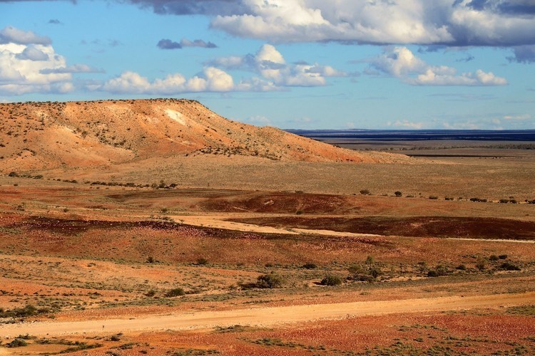 A rugged landscape with rolling hills and reddish dirt trails at Jump-up Loop in Sturt National Park, NSW, under a partly cloudy sky. The terrain is arid, with sparse vegetation.