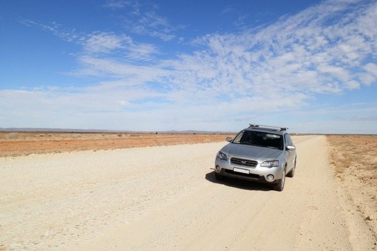 A silver SUV driving on the dusty, remote Strzelecki Track, surrounded by flat, arid landscape under a bright blue sky—one of the adventurous things to do in Copley, SA for outback explorers.