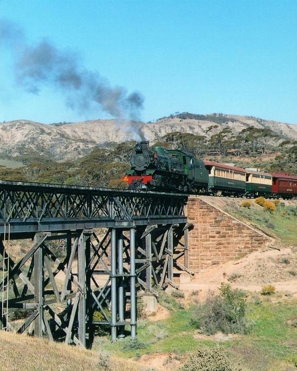 A steam train crosses an iron trestle bridge in the Flinders Ranges, with black smoke trailing behind, as part of a Flinders Ranges itinerary.