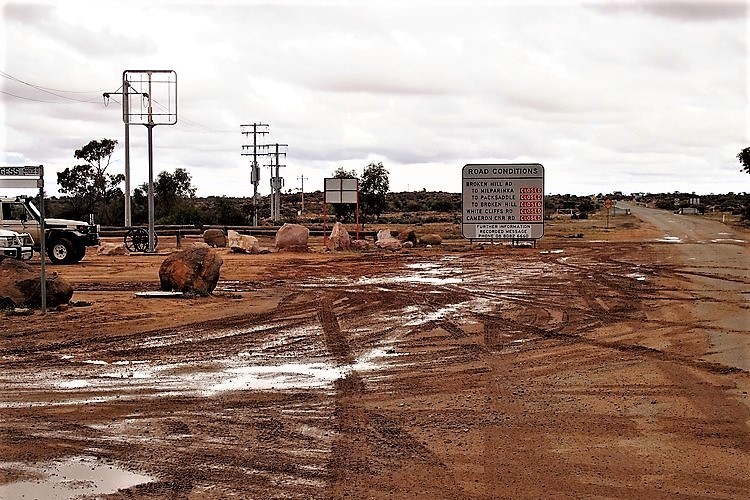 A muddy dirt road near Tibooburra, NSW, with large rocks and a sign displaying road conditions and closures, including the route to Cameron Corner, under an overcast sky.