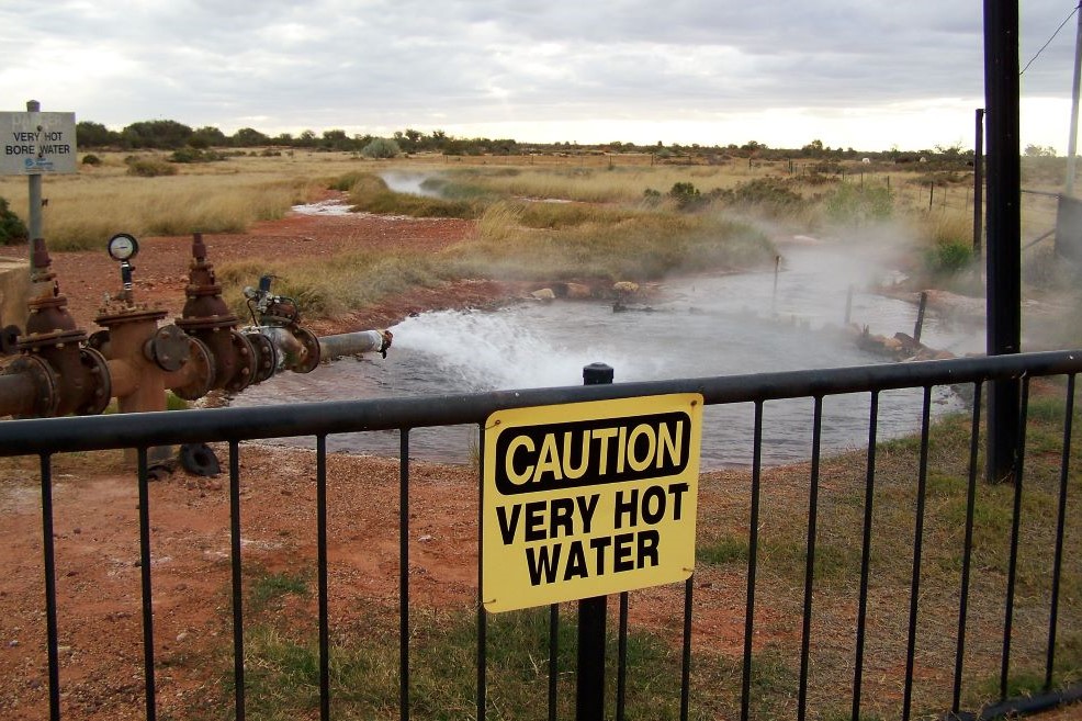 A "Caution Very Hot Water" sign at the Thargomindah bore, one of the interesting things to do in Thargomindah, with steam rising from the hot water and industrial pipes in an outback setting.