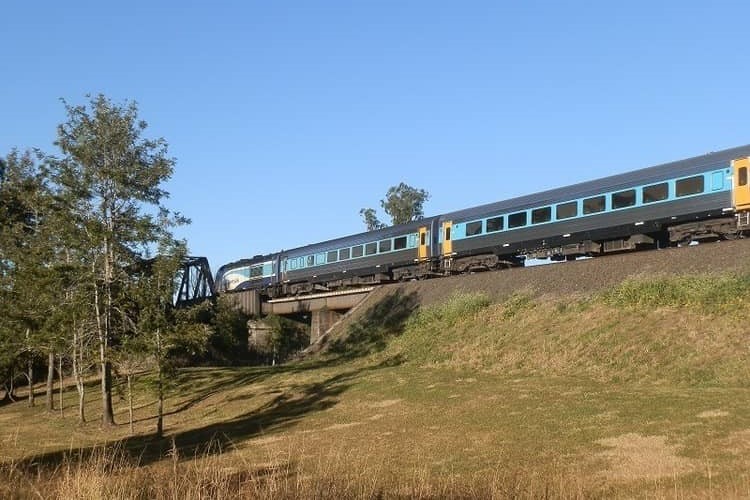 A passenger train crosses a bridge on Lions Road near Kyogle, surrounded by grassy fields and trees under a clear blue sky.