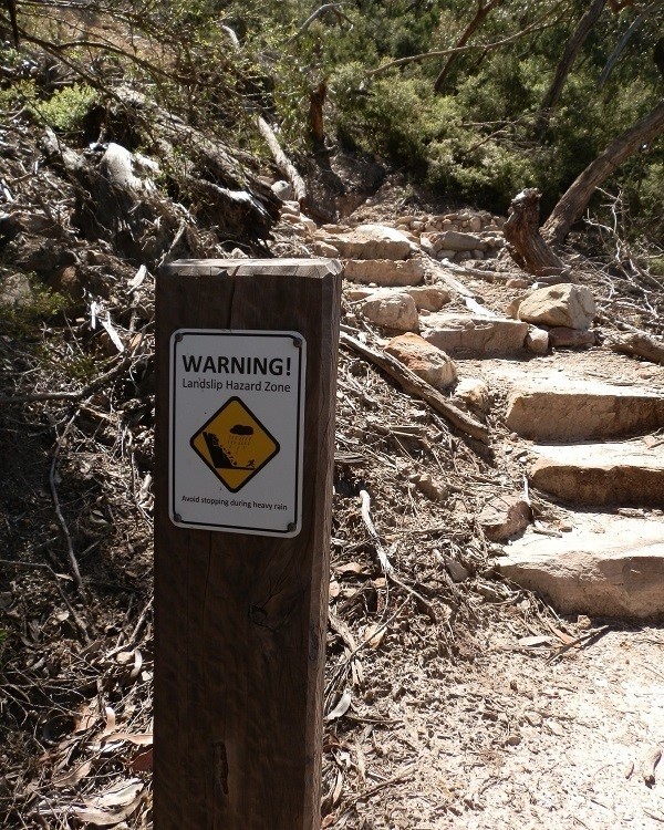 A "Warning! Landslip Hazard Zone" sign along the Mount Abrupt walk, advising hikers to avoid stopping during heavy rain. The sign is placed beside a rocky path surrounded by trees and dry foliage.