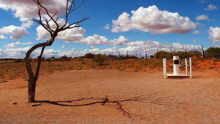 A barren tree casts a shadow on the dry, reddish dirt near a marker at Cameron Corner, NSW, under a bright blue sky dotted with fluffy white clouds.
