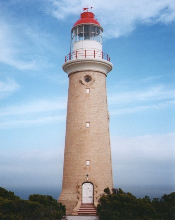 Cape du Couedic Lighthouse in South Australia, built with sandstone and featuring a red-topped lantern room. The lighthouse stands tall against a partly cloudy sky, surrounded by coastal vegetation.
