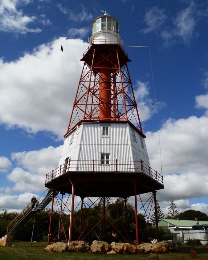Cape Jaffa Lighthouse, in Kingston SE, South Australia, features a unique iron structure with a red central column and white base. The lighthouse is elevated on stilts with a staircase leading up to the platform, set against a backdrop of blue sky and scattered clouds.
