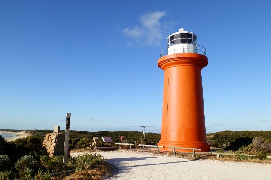 Cape Banks Lighthouse in Canunda National Park, Limestone Coast, South Australia. The vibrant orange lighthouse stands against a blue sky with a rugged coastal landscape and some nearby benches.