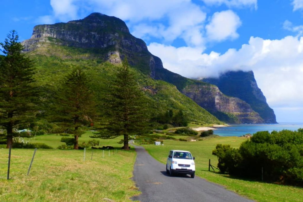 A small car drives along a road in the shadow of Mounts Gower and Lidgbird on Lord Howe Island, Australia. The towering green mountains rise in the background, while the limited car rental availability on the island makes exploring this scenic route a special experience.