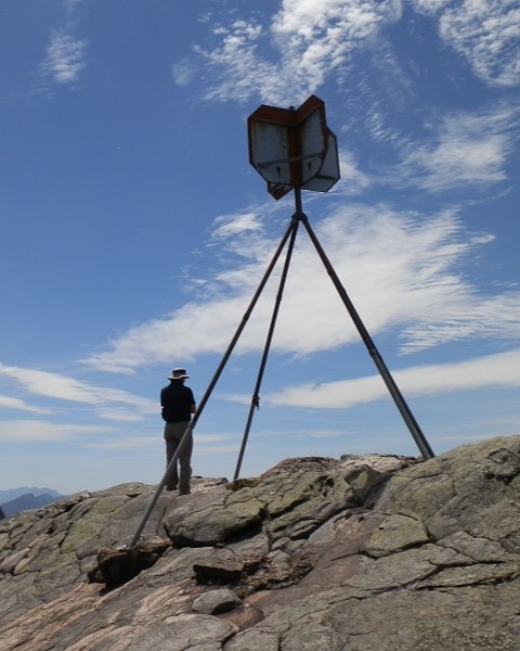 A hiker standing at the Mount Abrupt summit next to a metal trig point, against a backdrop of a clear sky with scattered clouds, capturing the achievement of the Mount Abrupt walk.
