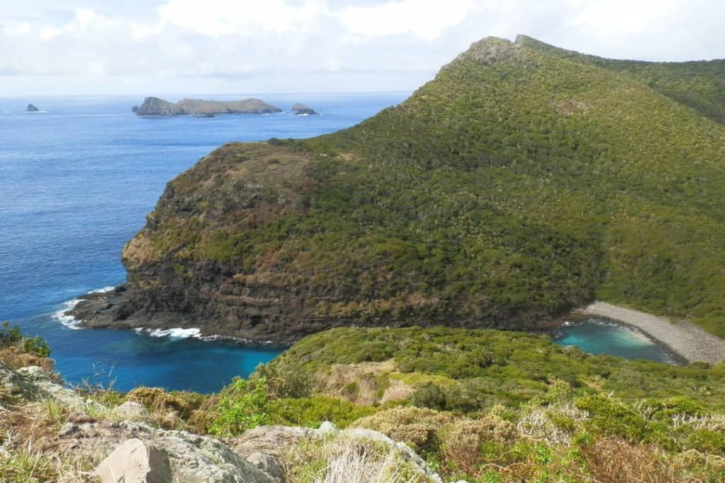 The view of Old Gulch from Mt Eliza on Lord Howe Island, Australia, shows a rocky coastline with green hills and a hidden cove with clear turquoise water.