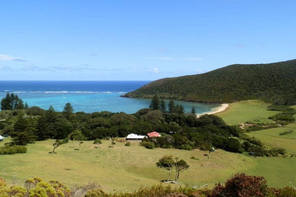 The view of Old Settlement Beach from Malabar Hill on Lord Howe Island shows a beautiful bay with clear turquoise waters, surrounded by green hills and trees. A few small buildings and a winding beach create a peaceful and scenic coastal landscape.