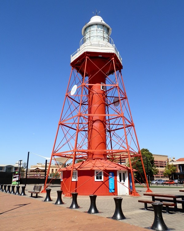 Port Adelaide Lighthouse, a striking red and white iron structure, stands in a public square. The lighthouse features a cylindrical tower supported by a lattice framework, with surrounding benches and urban structures in the background.