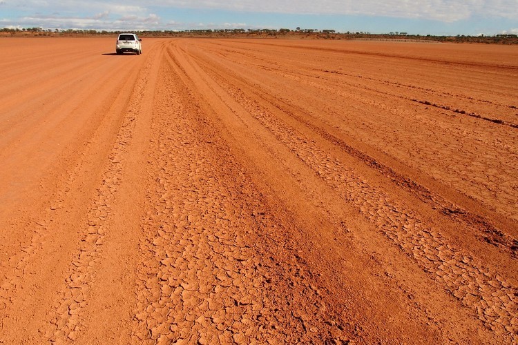 A lone vehicle drives on a vast, dry, and cracked red dirt road leading to Cameron Corner via Tibooburra.