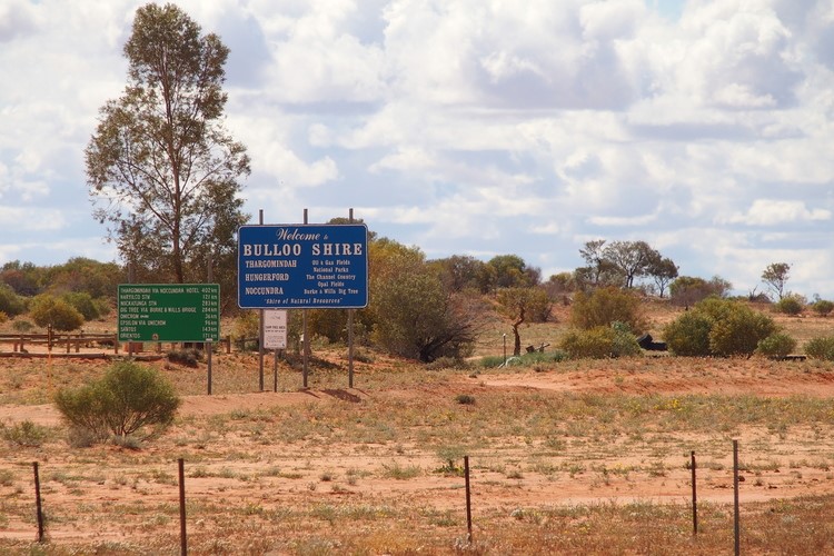 A "Welcome to Bulloo Shire" sign marking the entrance to the region seen against a typical Outback landscape of dry, sparse vegetation and a partly cloudy sky.