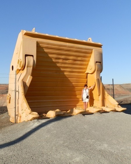 A person stands next to a massive yellow shovel bucket used in mining operations at the Super Pit Kalgoorlie, highlighting the enormous scale of the equipment used in the open-pit gold mine.
