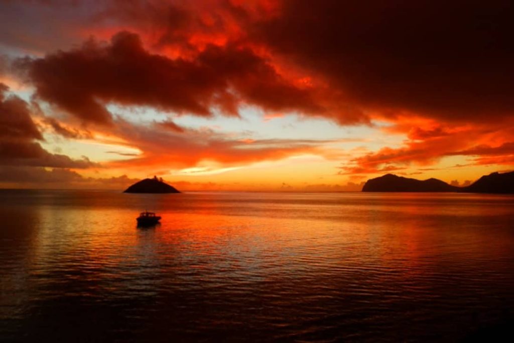 A vibrant sunset over the Lord Howe Island Lagoon, Australia, paints the sky in shades of red, orange, and gold. A small boat floats on the calm water, with the silhouettes of islands in the distance creating a stunning evening scene.
