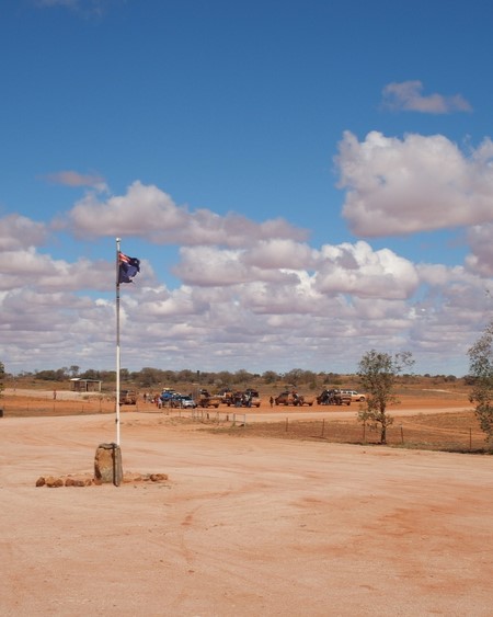 A distant view from the roadhouse to Cameron Corner, NSW, showing an Australian flag on a pole, dusty red terrain, and vehicles parked under a bright blue sky with scattered clouds.