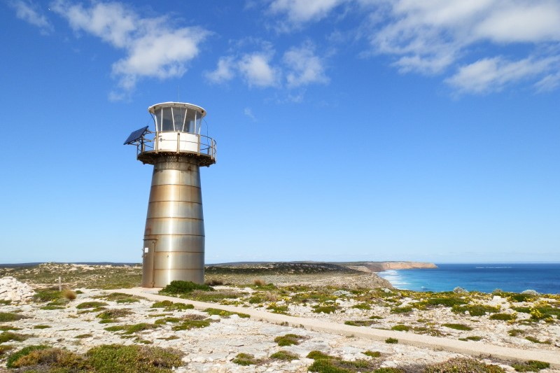 West Cape Lighthouse at Innes National Park, Yorke Peninsula, South Australia, situated on a rocky plateau with panoramic ocean views. The lighthouse is metallic in design and surrounded by wild coastal plants under a clear blue sky.