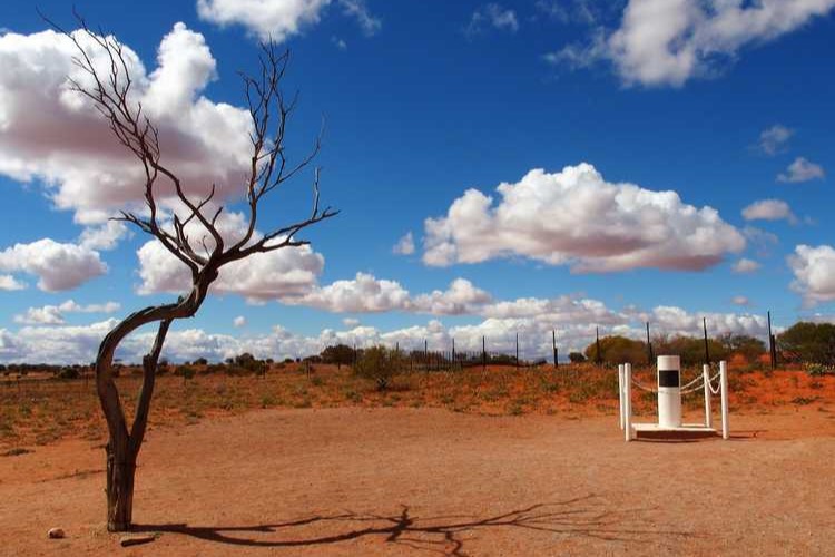 A marker at Cameron Corner, where three states meet, with a barren tree and vast outback under a sky filled with fluffy clouds, capturing a unique geographical spot in Australia.