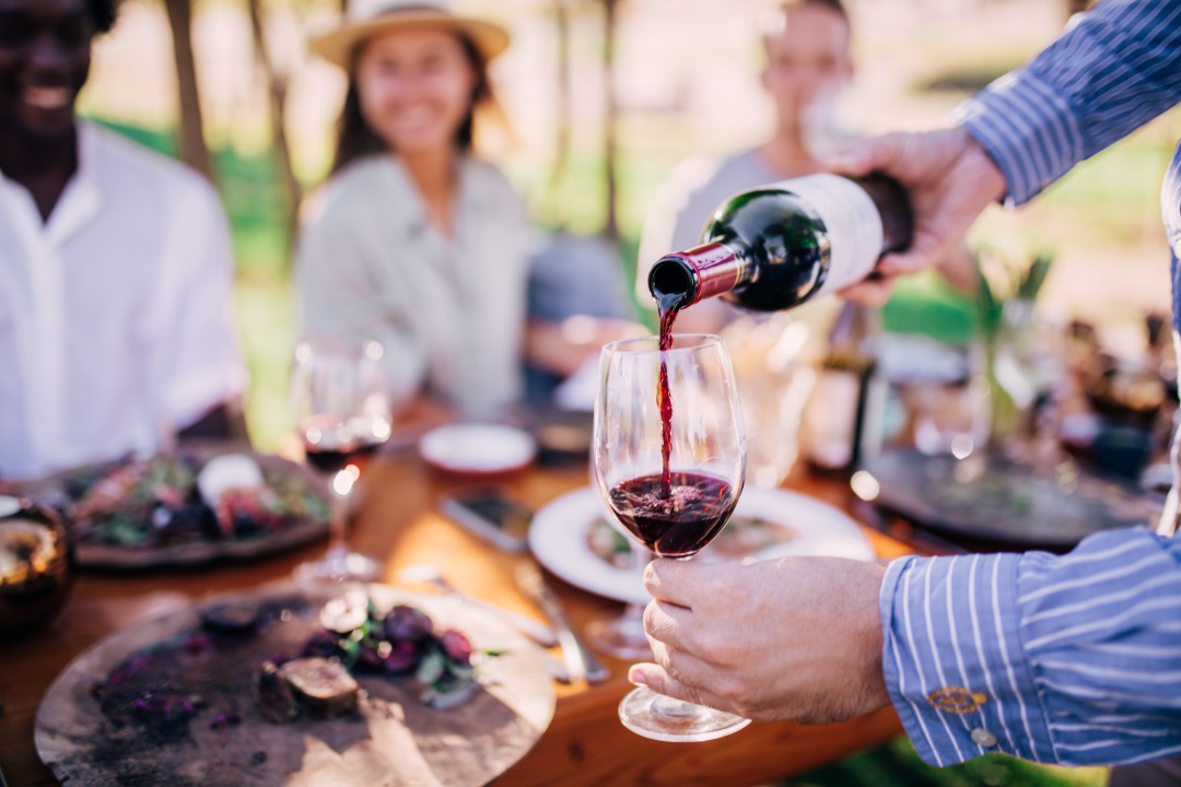 A glass of red wine being poured at an outdoor gathering, with blurred people and plates of food in the background.