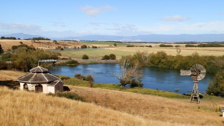 From Woolmers to Great Western Tiers, Tasmania, where there are several common Australian pests. A serene landscape featuring a small pavilion near a calm pond, with a rustic windmill and rolling fields extending towards distant mountain ranges under a clear blue sky.