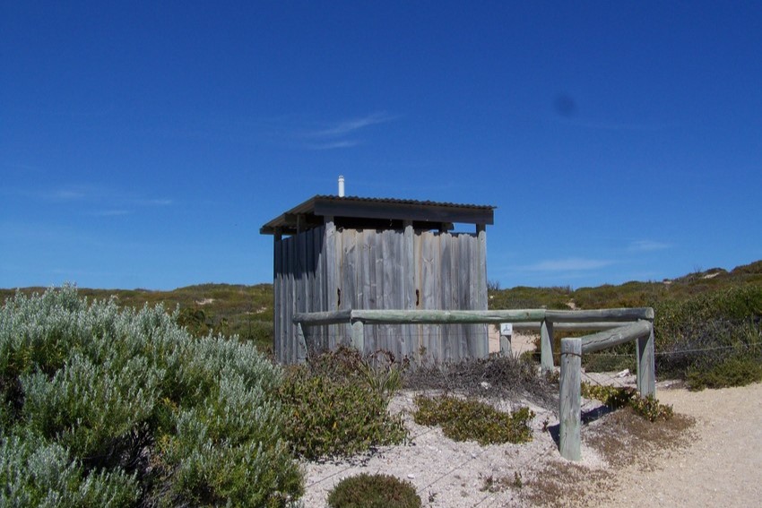 A rustic wooden public toilet facility located in the natural surroundings of Dhilba Guuranda-Innes National Park, Yorke Peninsula, South Australia, against a clear blue sky