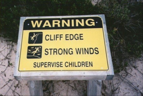 A yellow warning sign at Dhilba Guuranda-Innes National Park, Yorke Peninsula, South Australia, cautioning about strong winds, cliff edges, and child supervision.