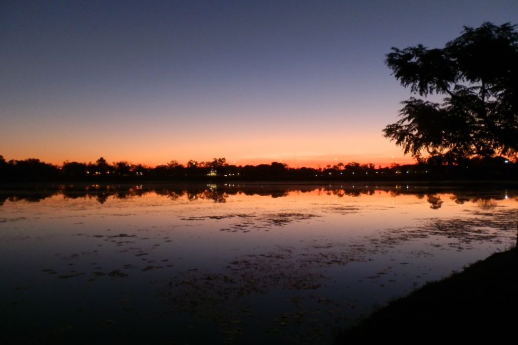 Tranquil scene of Kununurra Lake at sunset, with a vivid gradient sky of deep orange and purple hues reflecting on the water, surrounded by silhouettes of distant trees.