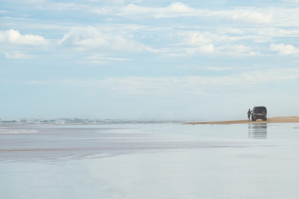 A person standing near a four-wheel-drive vehicle on a vast, empty beach with waves in the distance and a cloudy sky overhead, creating a sense of solitude and open space.