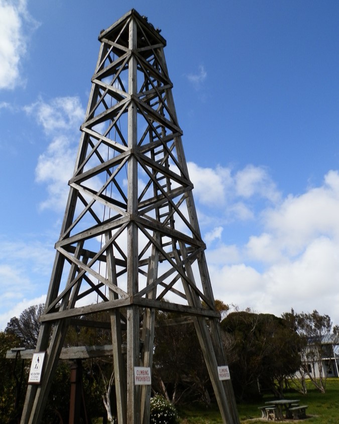 Historic wooden oil rig structure at Salt Creek, South Australia, standing tall against a blue sky with scattered clouds, surrounded by natural bushland.