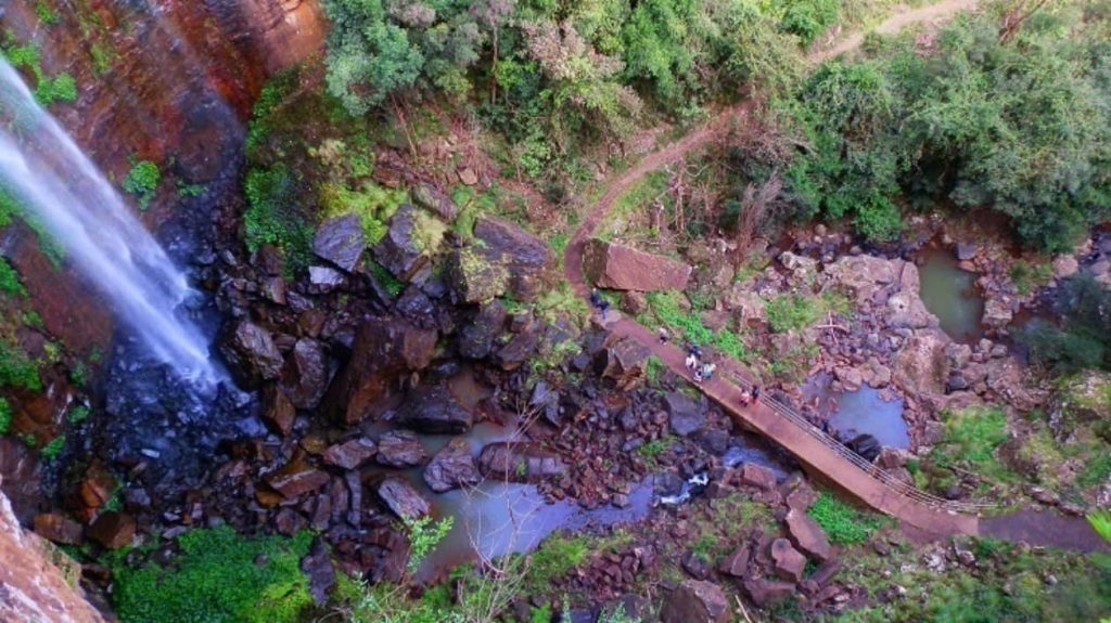 An aerial view of Queen Mary Falls with water cascading down rocky cliffs into a lush, green valley. A narrow walking bridge is visible below, where visitors are crossing and enjoying the scenery northwest of Woodenbong.