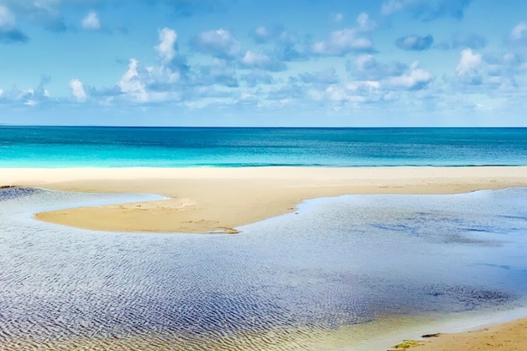 A serene beach scene with a calm, turquoise ocean stretching out to the horizon. A narrow Antechamber Bay, sandy spit separates the ocean from a shallow, tranquil lagoon, creating a picturesque and inviting landscape. The sky is a vibrant blue with fluffy white clouds.