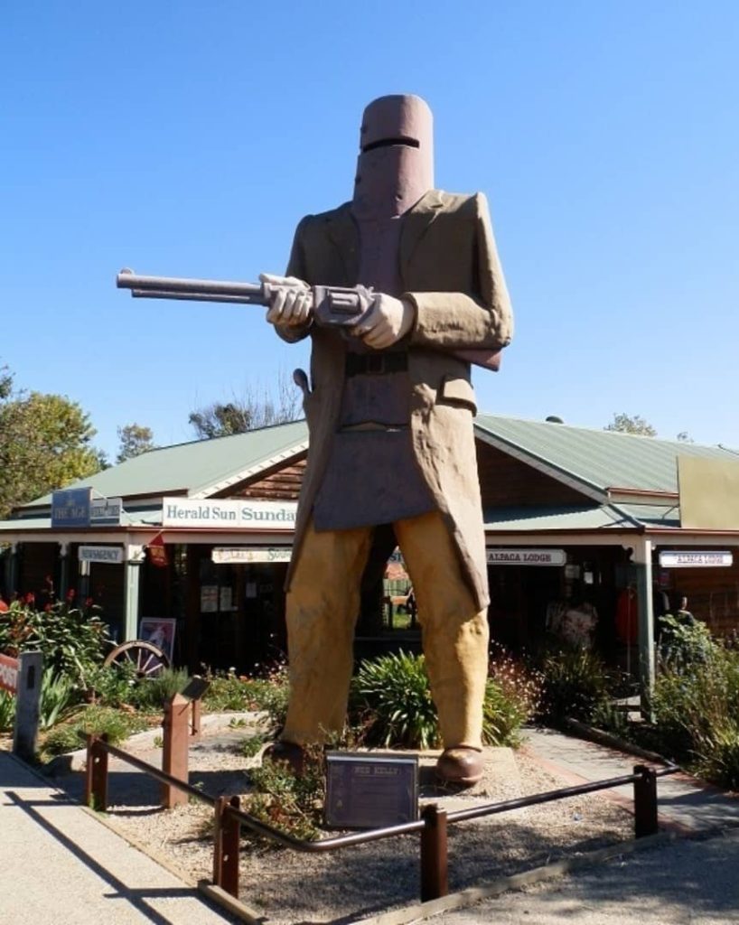 A large statue of a man wearing a helmet and holding a gun stands in front of a building. A woman stands in front of the statue, looking up at it.