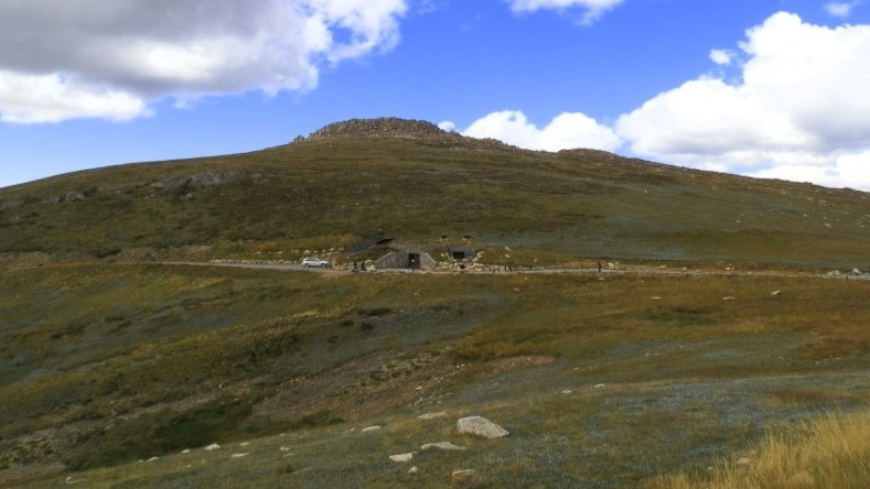 A grassy hillside with a small bunker-like structure near the Summit Road in New South Wales, set against a backdrop of blue sky and scattered clouds.