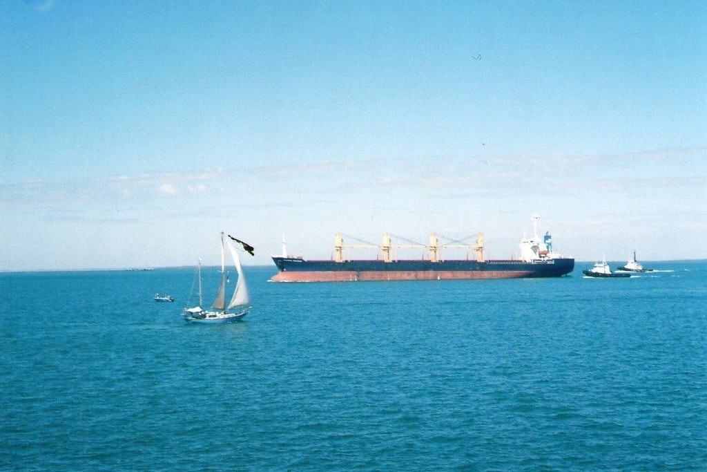 Darwin Harbour in the Northern Territory, Australia. A large cargo ship is anchored in the calm blue waters, accompanied by a few smaller vessels, including a sailboat and a tugboat. The expansive sea and clear sky create a peaceful maritime atmosphere, highlighting the harbor's significance as a busy port and sailing destination.