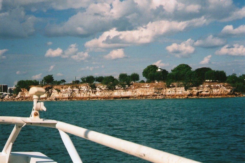 Darwin's cliffs as seen from the Mandorah Ferry. The rocky shoreline is lined with greenery and a few visible structures atop the cliffs, while the turquoise waters of the harbor stretch out in the foreground. The sky is partly cloudy, adding to the coastal scenery.