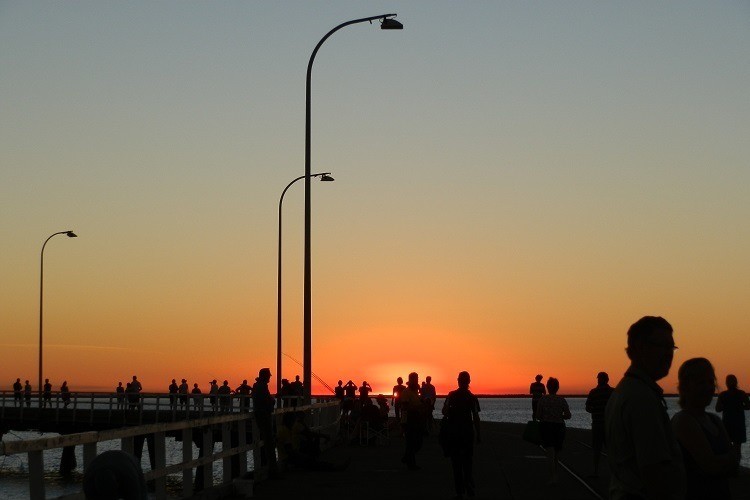 People silhouetted against a glowing orange sunset at Derby Jetty, Western Australia, with tall street lamps lining the pier—a peaceful activity and one of the scenic things to do in Derby WA.