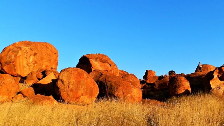 The image features the iconic Devils Marbles (Karlu Karlu) in the Northern Territory, Australia, showcasing large, rounded boulders scattered across a dry landscape. The rocks, glowing in warm orange hues from the sunlight, stand out against the bright blue sky. The scene captures the striking natural beauty and cultural significance of this unique geological formation.