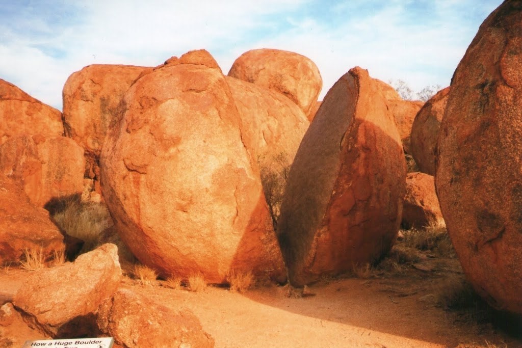 The image shows a close-up view of the Devils Marbles (Karlu Karlu) in the Northern Territory, Australia. It features large, rounded granite boulders, with one rock appearing split into two halves, showcasing the natural weathering and erosion processes. The red-orange hue of the rocks contrasts with the lighter sky, highlighting the unique geological characteristics of this iconic outback formation.