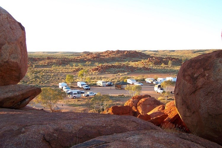 The Devils Marbles Campground in the Northern Territory, surrounded by a rugged landscape of large red boulders and sparse vegetation. Several caravans and campers are parked in the camping area, set against a backdrop of rolling hills and rock formations under a clear sky. The view is framed by massive boulders in the foreground, emphasizing the area's unique geological features.