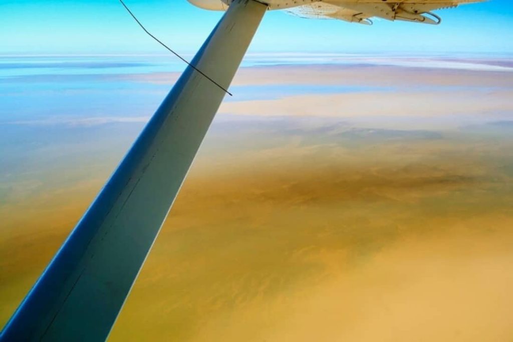 View from an aircraft flying over vast sand dunes in the South Australian outback, showing a mix of golden desert hues and blue sky.