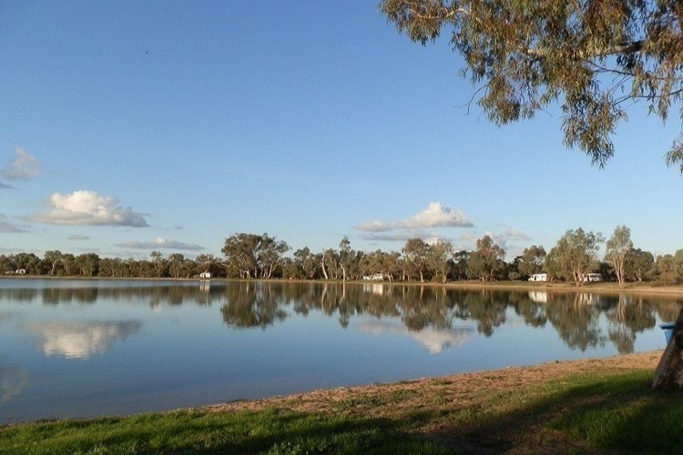 A calm, clear lake at Lake Lascelles, with trees lining the distant shore. The sky is blue with scattered clouds, and the water reflects the peaceful scene, showing a spacious free camping area.