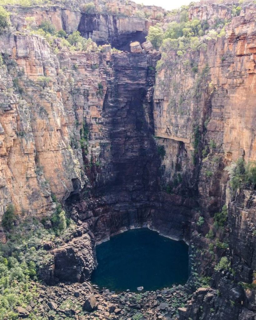 Jim Jim Falls in Kakadu National Park, Northern Territory, Australia. The towering rock cliffs surround a deep plunge pool, creating a dramatic and rugged landscape. The waterfall appears dry, with the dark rock face indicating where the water would normally flow during the wet season.