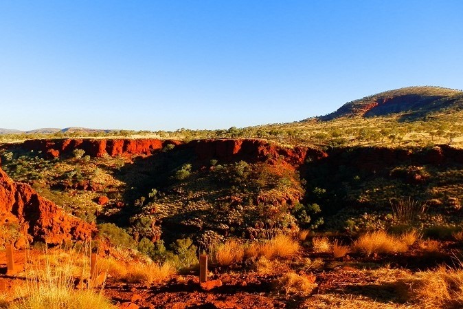 A scenic view of Karijini National Park in Western Australia, featuring rugged red cliffs and rolling hills under a bright, clear blue sky. Golden grasses and scattered green shrubs dot the foreground, adding contrast to the rich, earthy hues of the landscape.