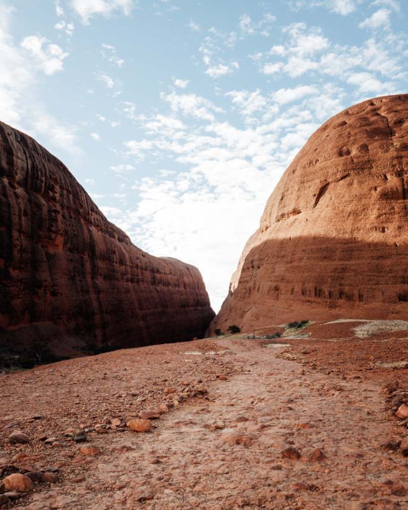 A rocky path leading between two massive, rounded rock formations at Kata Tjuta, also known as the Olgas, in the Uluru-Kata Tjuta National Park. The red earth and towering domes create a striking contrast with the bright sky, emphasizing the ancient and unique geological features of the area.