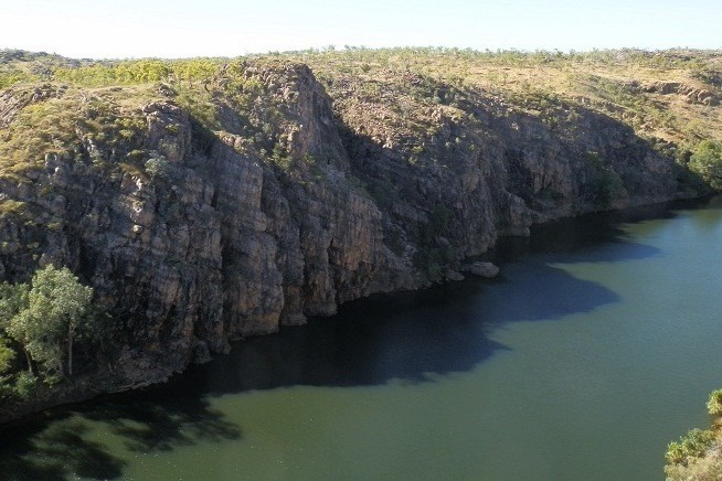The image depicts Nitmiluk National Park in the Northern Territory, Australia, featuring steep rocky cliffs that rise above the calm green waters of the Katherine River. The rugged terrain is covered with sparse vegetation, and the shadow of the cliffs falls over the water, creating a contrast between light and shade. The scene highlights the natural beauty and dramatic landscapes of this iconic outback location.