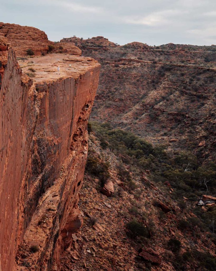 A dramatic cliff face of Kings Canyon in the Northern Territory, Australia. The rugged rock wall towers over the surrounding landscape, with sheer drops and layered textures highlighting the natural erosion. The expansive view captures the depth and scale of the canyon against a cloudy sky.
