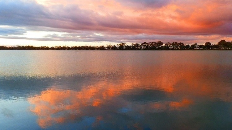 A dramatic sunset over Lake Lascelles with intense orange, pink, and purple clouds reflected in the lake's still water, contrasting with the dark silhouette of trees on the horizon.
