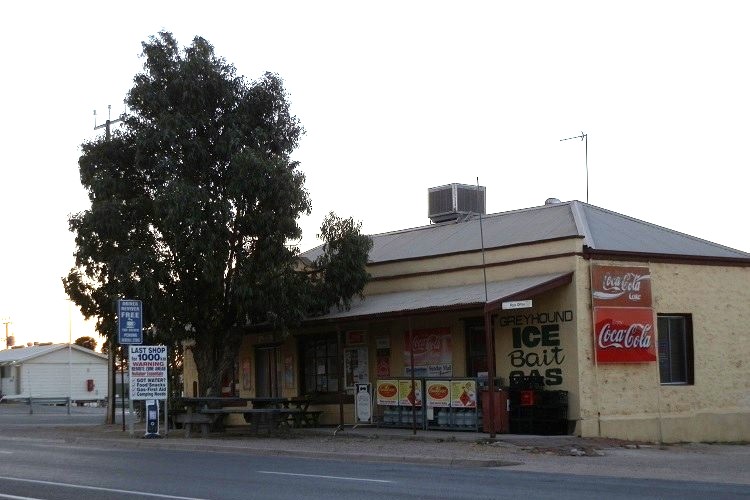 Front view of the last shop in Penong, South Australia, offering essentials like ice, bait, and gas, with Coca-Cola signs and seating outside.