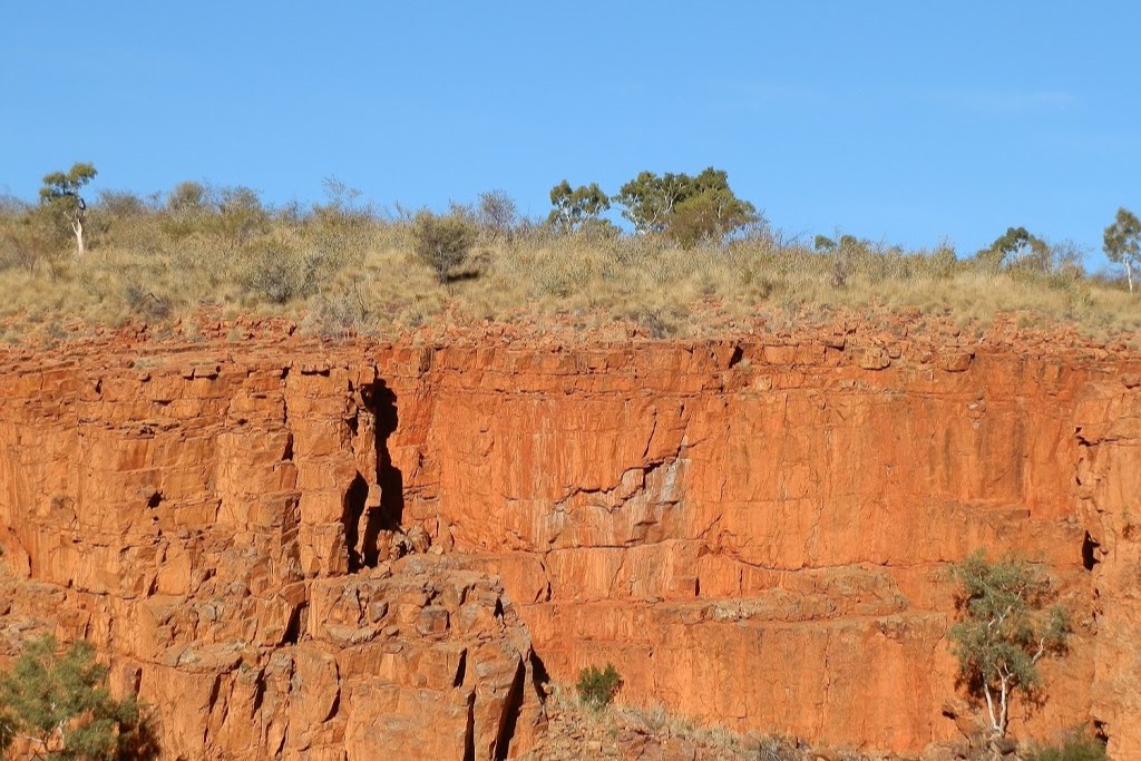 The red cliffs of Ormiston Gorge illuminated in late afternoon light, highlighting the rugged textures, a scenic stop among things to do in Central Australia. Sparse greenery tops the cliff under a clear sky.